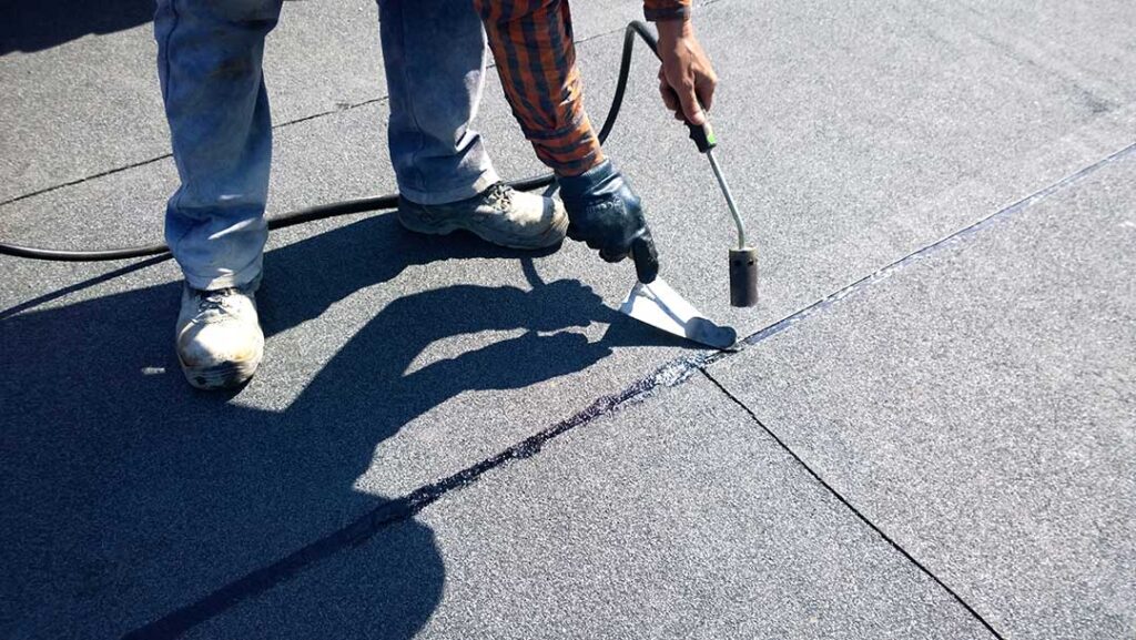 A homeowner examines aging shingles and roof flashing beside a snow-covered residential roof.