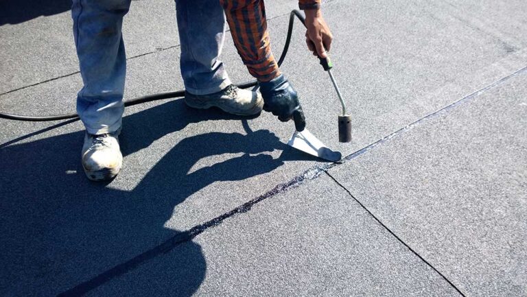 A homeowner examines aging shingles and roof flashing beside a snow-covered residential roof.